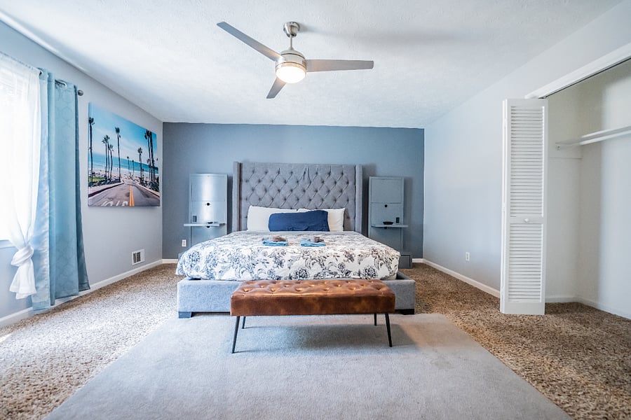 Spacious primary bedroom with a gray tufted headboard, a blue accent wall, a wooden bench, and ample closet space.