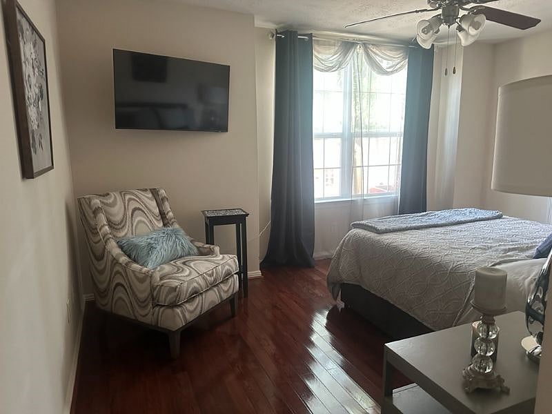 Additional view of the guest bedroom with cozy seating by the window and blackout curtains paired with sheers, providing both privacy and flexible lighting. The room offers a peaceful, comfortable retreat with hardwood flooring and plenty of space.