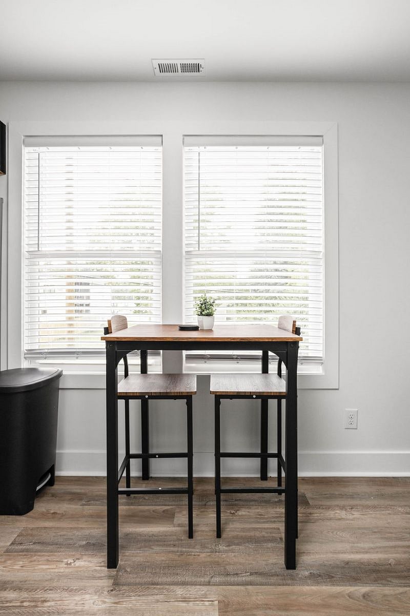 Cozy dining nook with a rustic wooden table, metal-frame stools, and bright natural light from large windows.