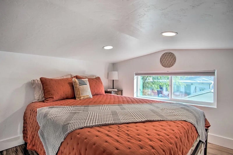 Queen size super comfy bed in the loft space with a blackout shades, nightstand with lamp and a hanging rack for clothes. There are some storage boxes beneath the bed for additional storage. 