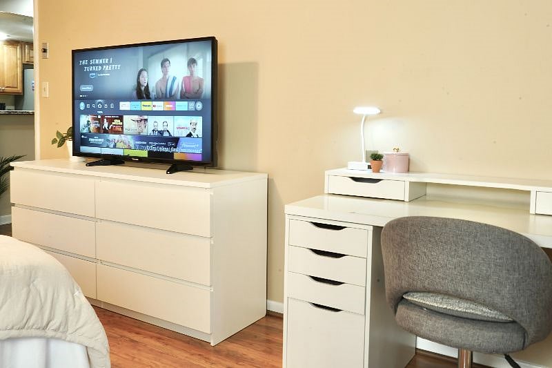Very spacious primary bedroom with queen memory foam bed, vanity, desk, dresser, TV, walk-in closet, and en suite bathroom. Double french doors with view and access to enclosed courtyard.