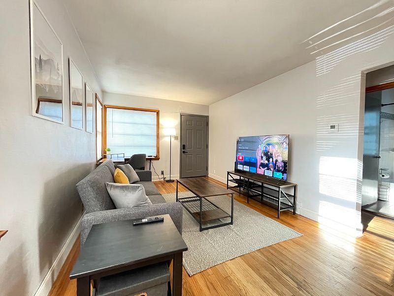 Living room with desk work space and chair, art work, coffee table, end table, tv stand, 58" TCL Google Smart TV, rug, lamps, usb charger and outlet extender, 500mbps WiFi internet, queen blanket, throw pillows, foldable futon couch for additional full-sized sleeping space, and dvd player.