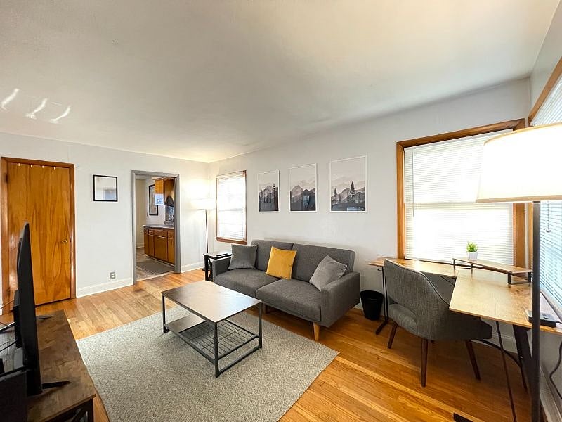 Living room with desk work space and chair, art work, coffee table, end table, tv stand, 58" TCL Google Smart TV, rug, lamps, usb charger and outlet extender, 500mbps WiFi internet, queen blanket, throw pillows, foldable futon couch for additional full-sized sleeping space, and dvd player.