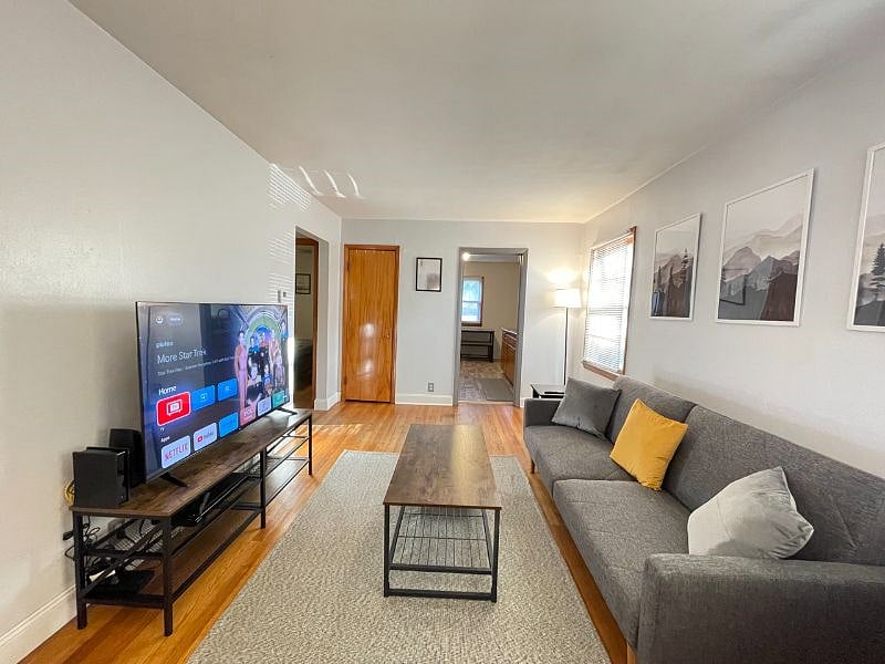 Living room with desk work space and chair, art work, coffee table, end table, tv stand, 58" TCL Google Smart TV, rug, lamps, usb charger and outlet extender, 500mbps WiFi internet, queen blanket, throw pillows, foldable futon couch for additional full-sized sleeping space, and dvd player.