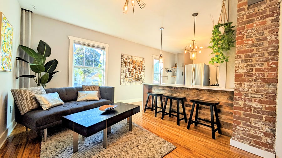 The living room has modern furniture, Smart TV, bar seating at the kitchen and unique exposed brick