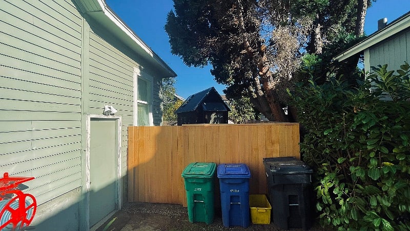 The trash and recycling bins just outside the gate / fence in the driveway. 