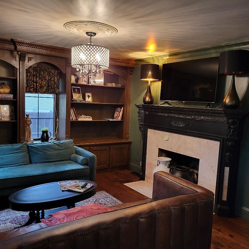 Living room with two couches, one leather and other green suede. Dark moody library with plenty of windows, fireplace, books, games and antiques for the decor.