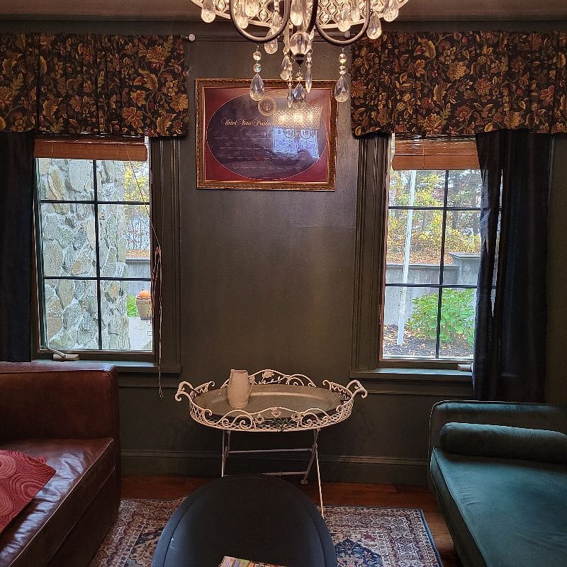 Living room with two couches, one leather and other green suede. Dark moody library with plenty of windows, fireplace, books, games and antiques for the decor.