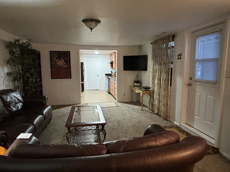 Living room is carpeted and has a leather couch, a loveseat, and a 42" smart television. Entry to apartment is pictured on the right.