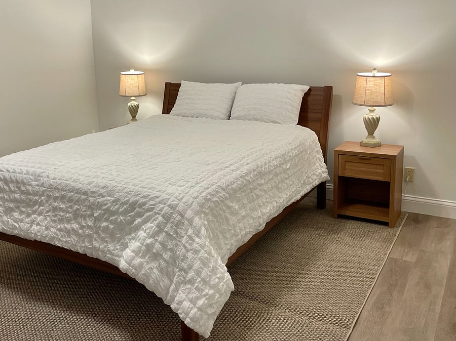 Bedroom with soft sheets and comforter, side tables and lamps. LVP flooring and area rug.