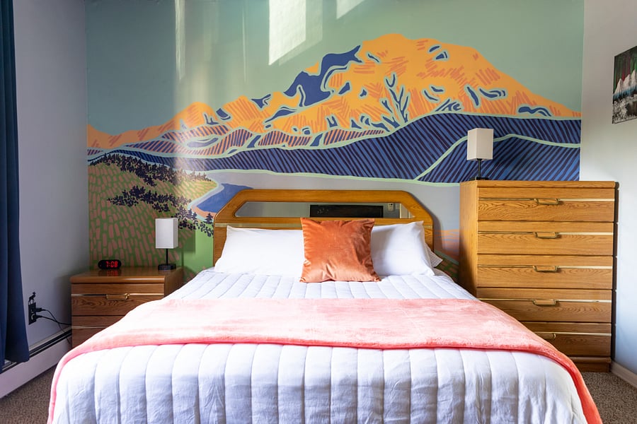The Denali Room with queen bed, ceiling and portable fans, extra pillows and blankets, and portable heater. The wall mural is a hand-painted depiction of Denali (The Great One), previously known as Mount McKinley.