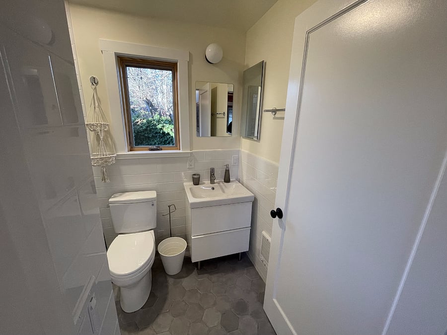 Bathroom facing in from bathroom door. Two mirrors, toilet, 2-drawer bathroom vanity, and side-facing window.