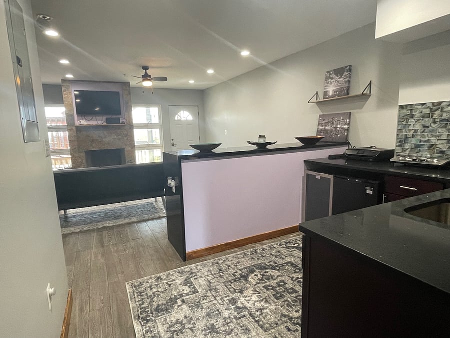 Photo of kitchenette and living room which features lots of sunlight, wood burning fireplace and wall-mounted smart TV.