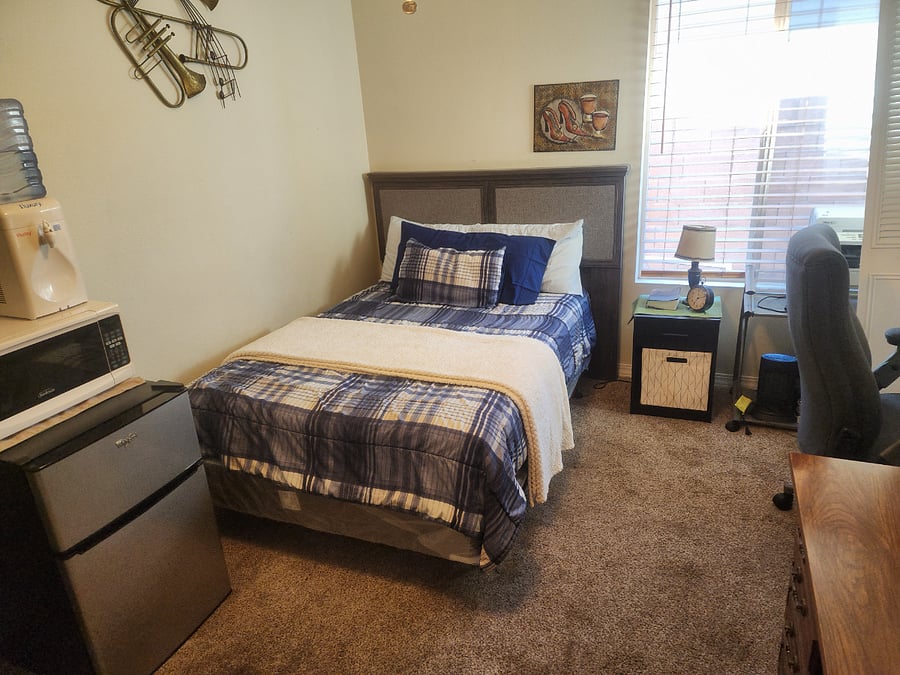 Twin bed, nightstand with nightlite. Fridge/freezer, microwave oven,  and water cooler also seen. 