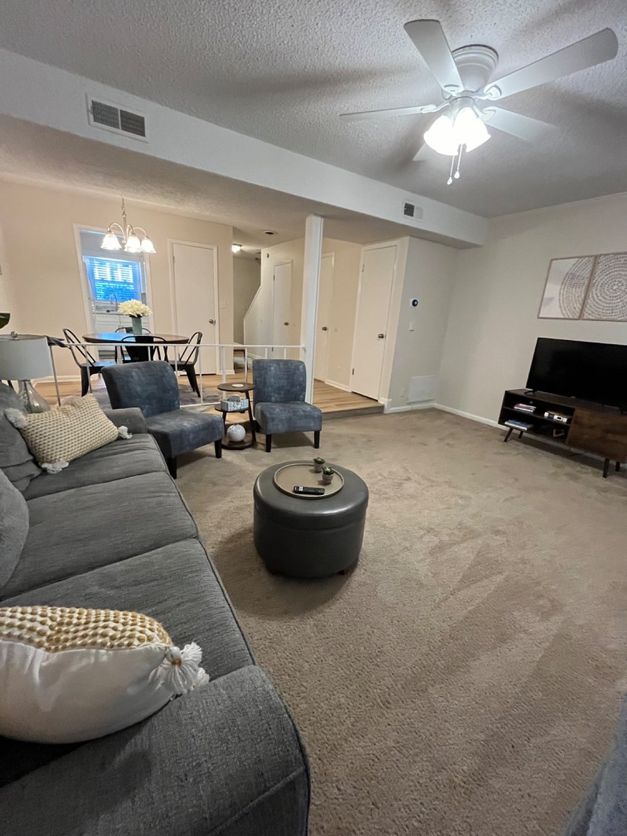 Living room with couch and two chairs, a footstool, and a smart TV. There are also games in the footstool as well as books and cookbooks under the TV. There is a wireless printer on the bookshelf as well.