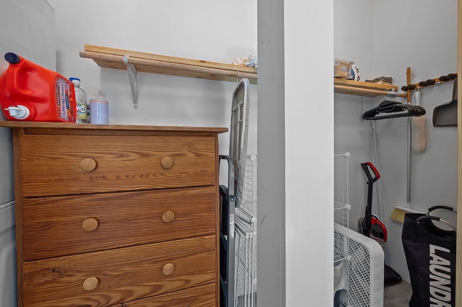 The closet interior includes a dresser, laundry essentials, compact ironing board & iron, hangers, basic cleaning equipment including a vacuum, basic cleaning supplies, laundry hamper & basket, box fan, & extra storage shelves.