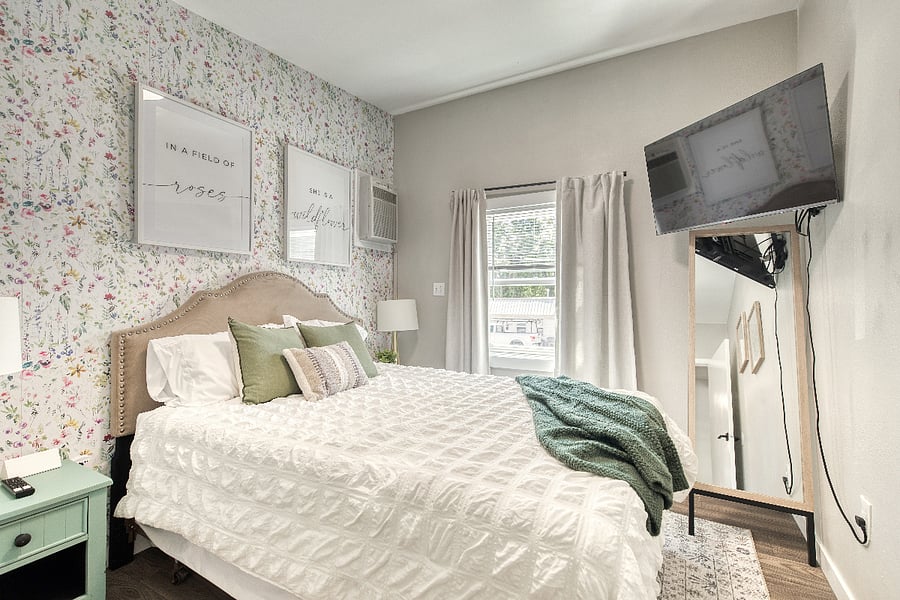 The Wildflower bedroom, located behind the mountain mural, has a queen bed, TV, floor mirror, two nightstands with lamps, and a closet (not pictured) for guests to relax and feel at home in.