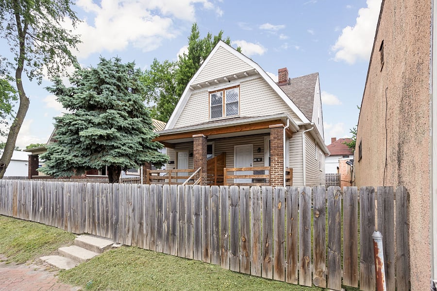 Charming two-story home with a welcoming front porch, wooden fence, and lush greenery.