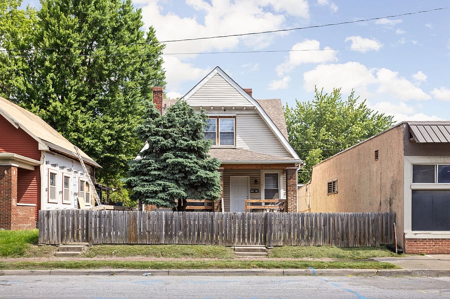 A charming two-story home with a fenced front yard, a covered porch, and a large tree, situated between commercial and residential buildings.