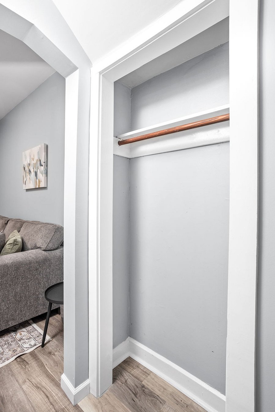 Small open closet with a wooden hanging rod, adjacent to a cozy living space with a gray couch and decorative pillows.