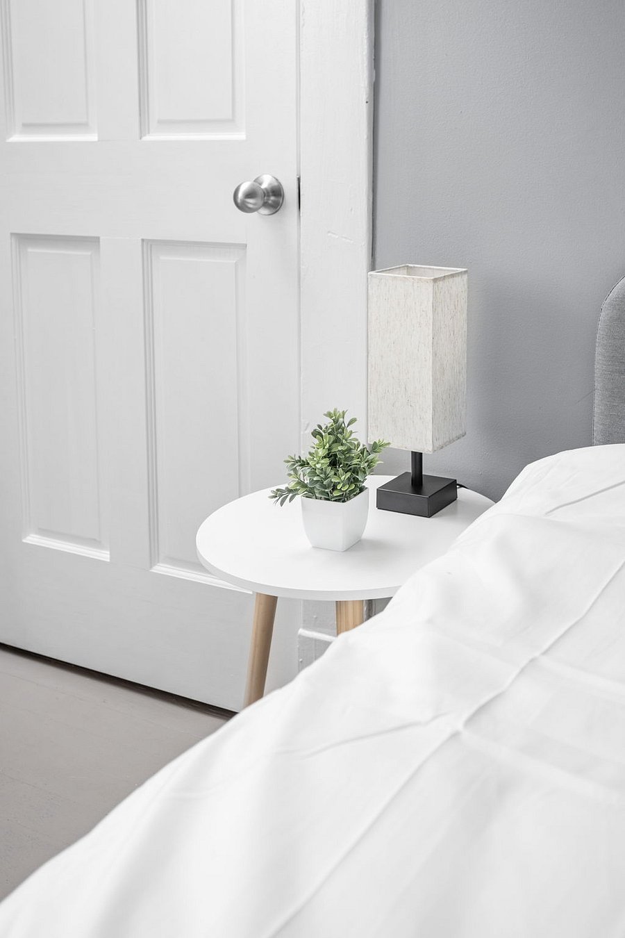 Minimalist bedside setup featuring a white round table, a small potted plant, and a modern lamp next to a neatly made bed.