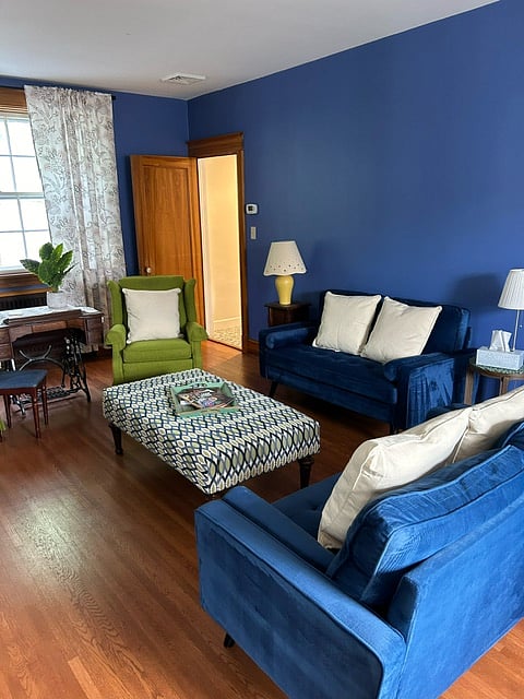 The living room is a serene space filled with blue and green hues, two love seats, arm chairs, an oversized ottoman, wall art, a large work desk, fireplace (non-functioning), and TV.