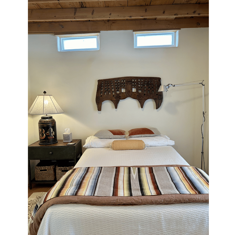 Full-size Temperpedic mattress with organic cotton sheets, Belgian linen duvet, vintage saltillo throw. Dual bedside lighting. Clerestory windows above bed under wooden beam ceiling.
