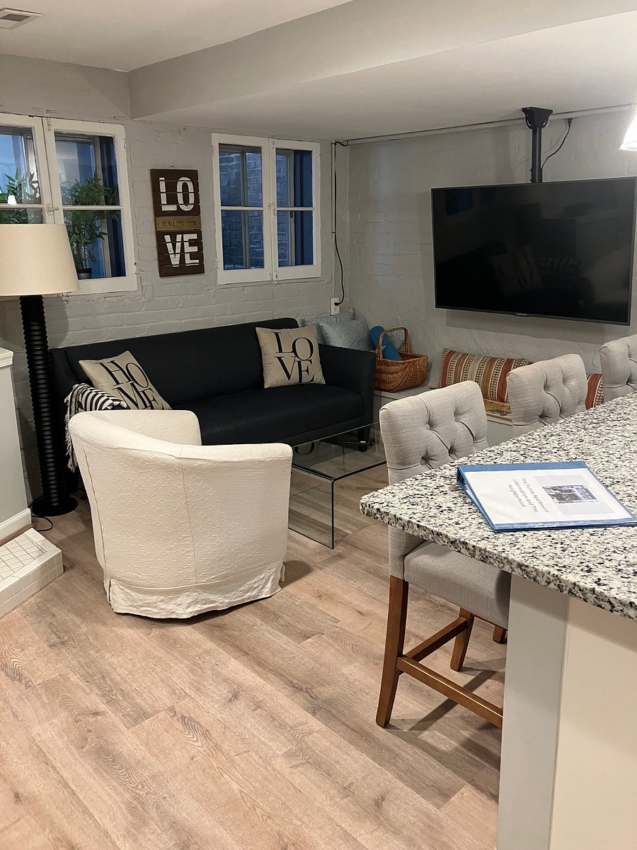 Living area with wall-mounted TV, couch and armchair, plus 3 barstools at the countertop connected to the Kitchen area.