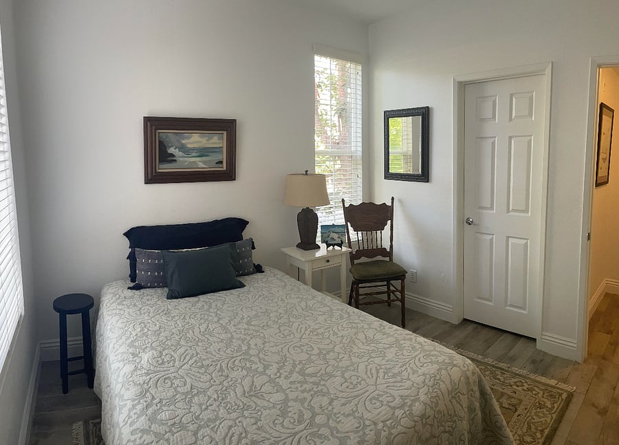 Room includes a full/double bed, wool rug, 2 chairs, throw pillows, a side table and lamp, a bookcase, mirror, and artwork. The walk-in closet is located to the right of the chair.