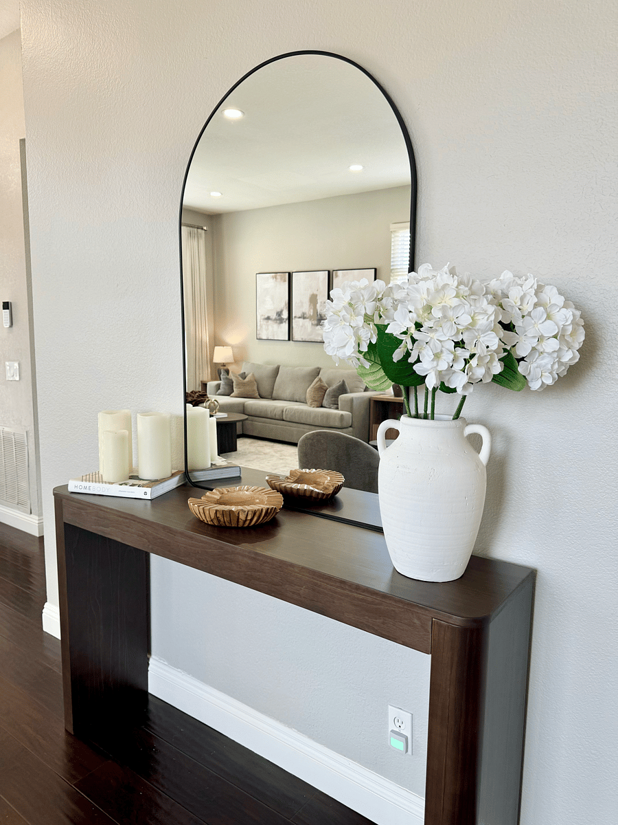 An elegant entry console with a sleek design is paired with a wall-mounted mirror above, creating a stylish focal point. The console features a small dish to conveniently hold keys & other small essentials, keeping the space organized.