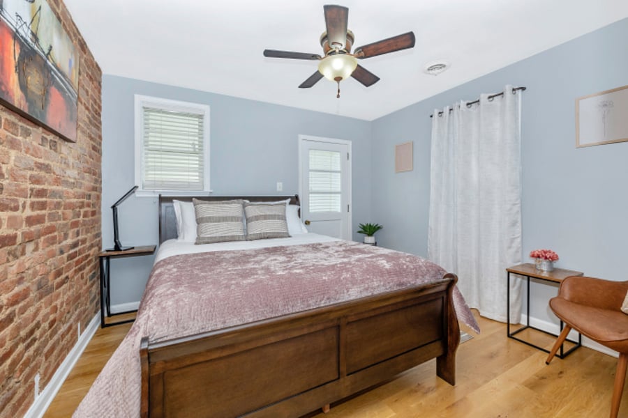 Primary bedroom with room darkening shades, ceiling fan, dresser and exposed brick walls.