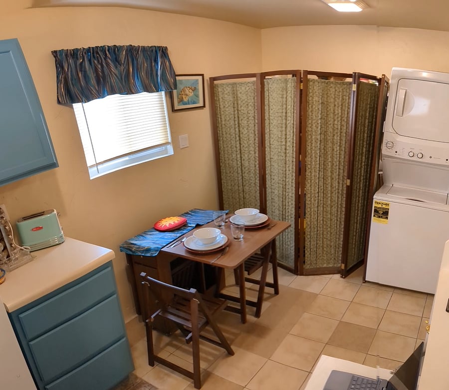 edge of kitchen, shows toaster, dining table folds down for more room, folds out to seat four. chairs are stored inside unit, shelving behind screen, stacked washer dryer unit