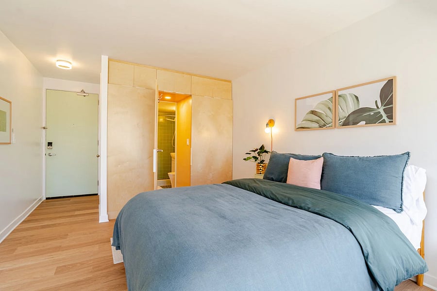 Sleeping area with custom birch cabinetry, wall sconce, and bright sliding glass door view of the entry and concealed bathroom door.