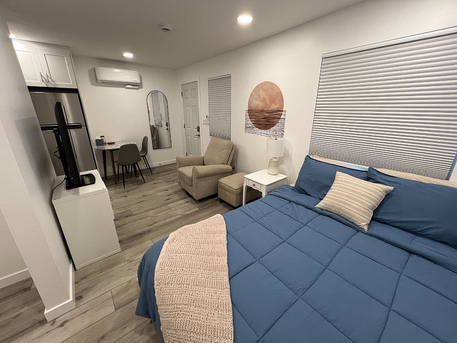 View from the sleeping area. Comfy queen size bed, room darkening shades, lounge chair and ottoman, dining table with chairs, and full size mirror.