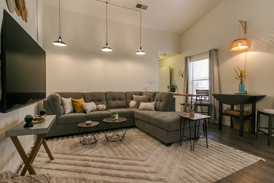 This living room space is designed with comfort and modern style in mind, creating a cozy and welcoming environment. Key elements include:
1. Sectional Sofa: A large, plush gray sectional sofa with a variety of throw pillows, offering ample seating and inviting relaxation. This setup is ideal for groups or families to lounge together comfortably.
2. Coffee Tables: Three round coffee tables with metal legs and wooden tops provide a central gathering space for drinks, books, or decor items. Their varied heights and compact size add visual interest and flexibility in use.
3. Rug: A soft, textured rug defines the seating area, adding warmth and comfort underfoot. Its subtle design complements the rooms modern decor.
4. Accent Tables: Side tables with natural wood and metal elements enhance the modern rustic vibe, holding small decorative items and greenery to add a natural touch.
5. Lighting and Ambiance: The room has layered lighting, with pendant lights casting a soft glow and natural light streaming through the window. The placement of lamps and wall fixtures ensures a balanced ambiance suitable for both day and night.
6. Open Layout: The space flows seamlessly into the adjacent dining area, promoting an open and airy feel ideal for socializing or relaxation.
This living room is well-suited for tenants looking for a stylish, spacious, and comfortable space to unwind, with a layout that accommodates both small and larger groups effectively.