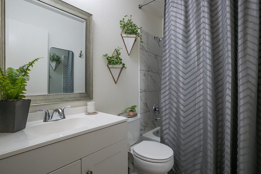 Unwind in this thoughtfully designed bathroom, featuring a modern vanity with ample storage and a sleek silver-framed mirror. Soft lighting complements the minimalist decor, with fresh green accents from hanging plants that add a natural touch. The shower area boasts a stylish, chevron-patterned curtain alongside elegant tile work, creating a spa-like atmosphere. This bathroom combines functionality and aesthetics, providing a refreshing space for your daily routine.