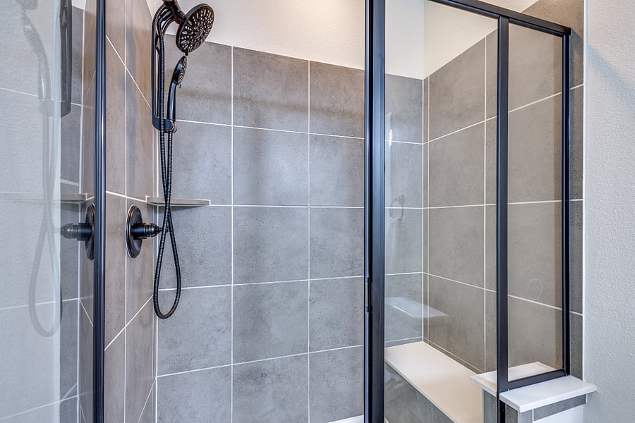 Walk in modern shower with walls decorated with tile that includes shower head, handheld shower faucet, shelf with shampoo/conditioner/body soap wash, and full bench seating built into the wall.