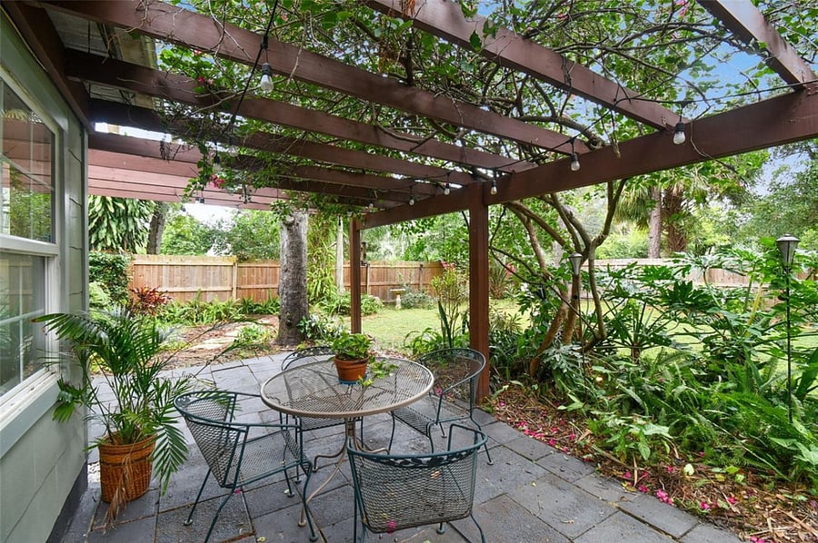 The paver patio features bistro-style seating perfect for morning coffee or al fresco dining. Mature trees provide natural shade while flowering vines and string lights create an intimate ambiance. Privacy fencing and established gardens with palms, ferns, and colorful plantings complete this serene Florida oasis.