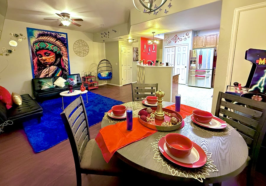 This shared area is vibrant and eclectic with bold decor, including a colorful mural & hanging chair. The dining area has a wooden table set with red dishware, candles, & a gold centerpiece.