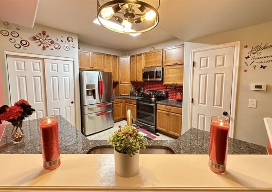 This kitchen showcases warm wood cabinets, dark granite counters, and a bold red tile backsplash with stainless steel appliances. Decorative elements and plants add charm and warmth to the spa