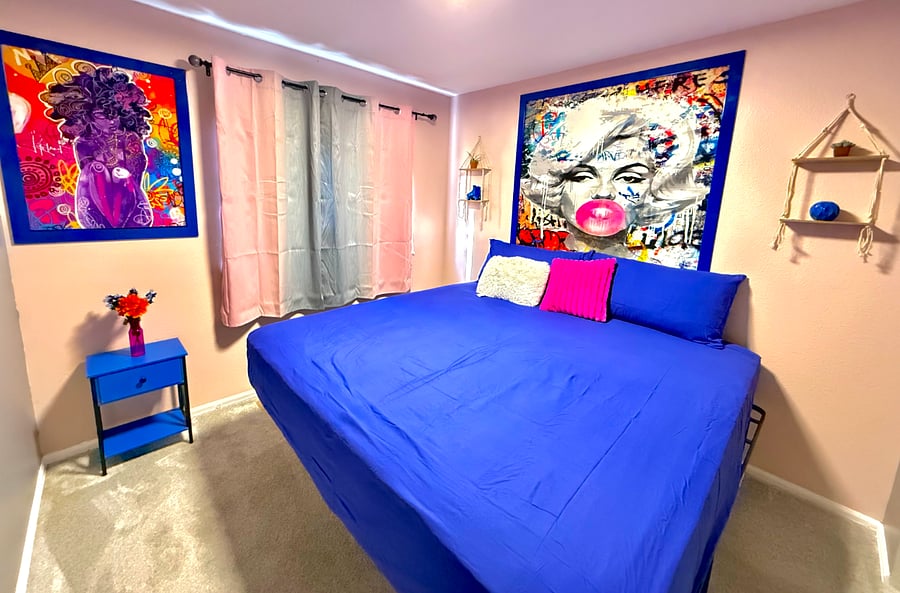 The bedroom exudes vibrant & eclectic energy with its bold, colorful artwork & furnishings. The large bed, adorned with deep blue linens, invites relaxation, while the bright artwork adds a playful touch. Natural light streams through the window.