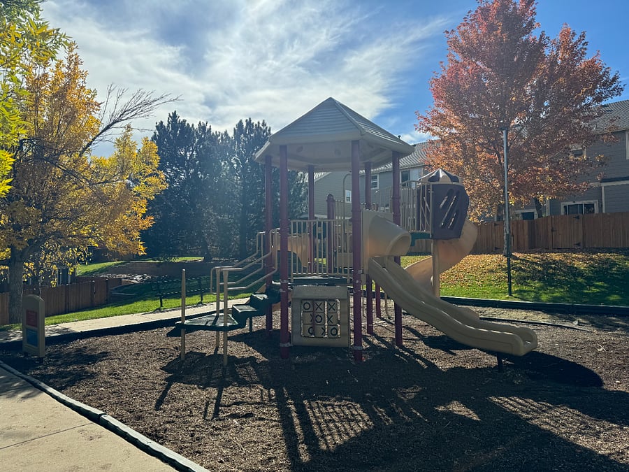 A vibrant playground bathed in sunlight, surrounded by refreshing trees and lush green grass. This inviting space features a cozy sitting area and a charming playhouse w/ slides, perfect for children to enjoy hours of fun & adventure in the great outdoor