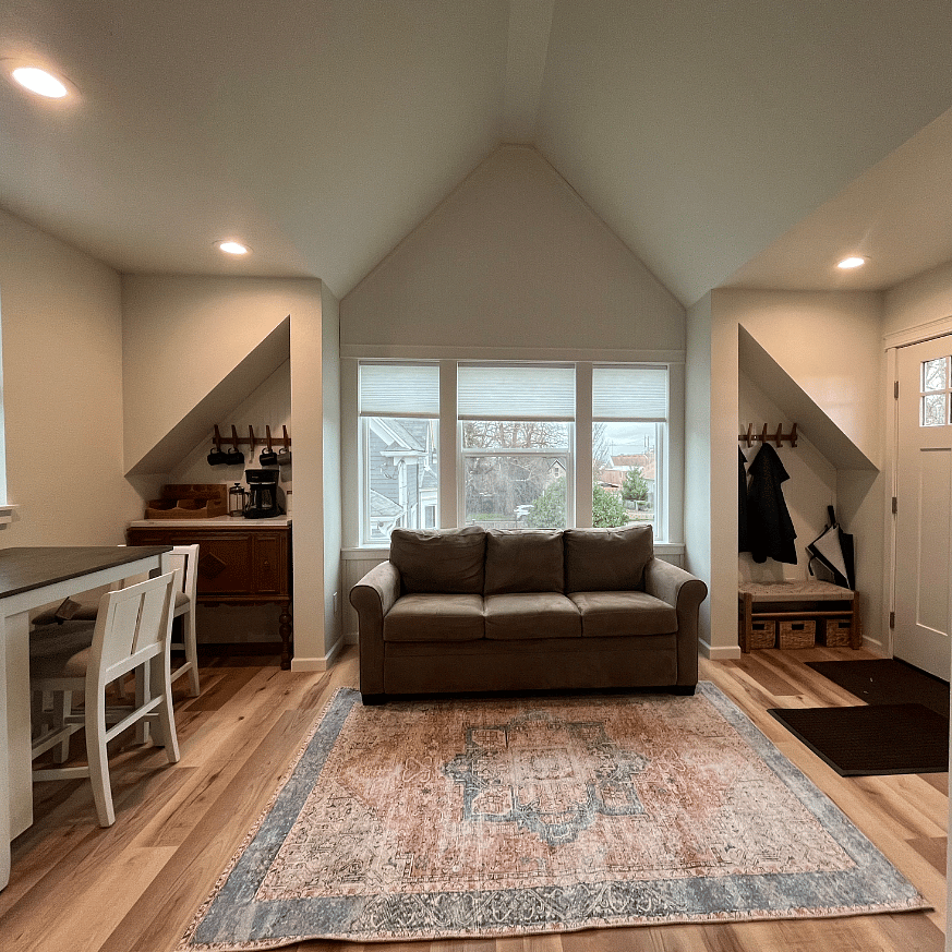 Open living space with queen hide-a-bed couch, entry, coffee bar, eat-in area, and kitchen. Large windows overlook yard, tree tops and gardens.