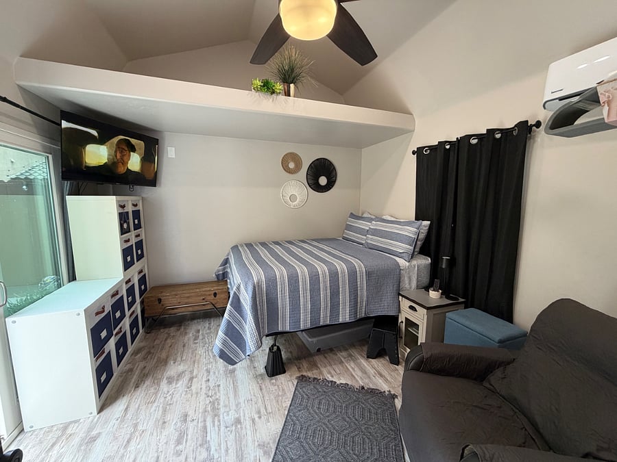 Bedroom with queen-size bed, smart TV, including e-recliner, e-night stand, shelving with hanging rods for clothing and sliding glass door to exterior. 