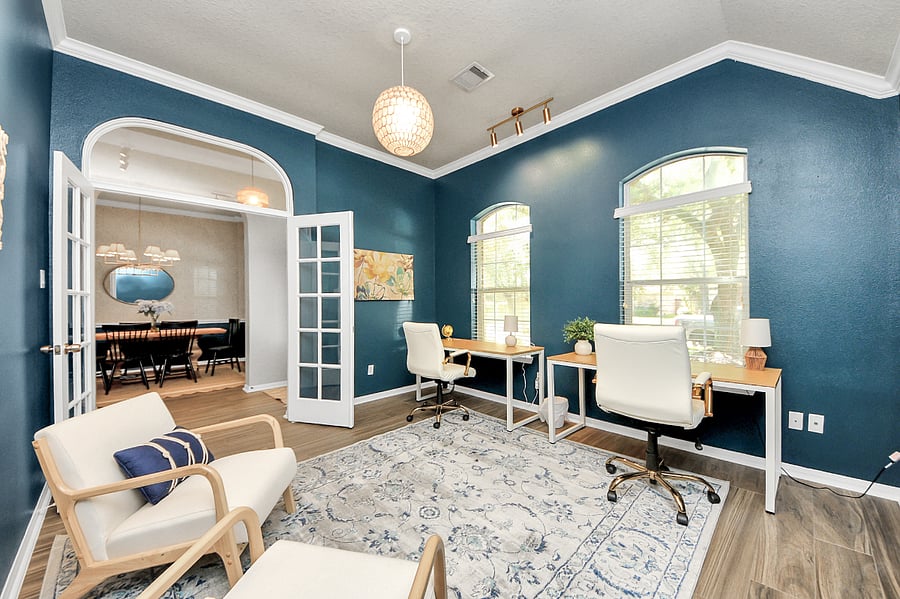 Spacious office with fresh paint, wood plank tile flooring, and French doors for a touch of elegance. Features dual desks with a comfortable sitting area, accented by modern track lighting and a stunning capiz orb pendant light.