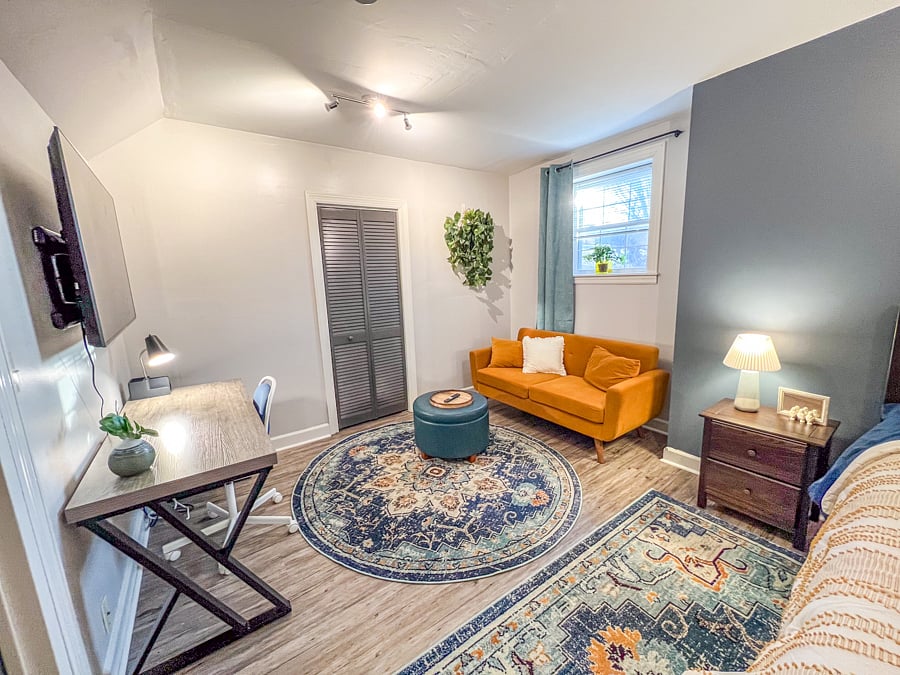 Large bedroom with wall-mounted smart tv, couch, ottoman, work desk and chair.
