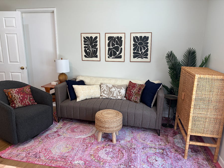 warm, inviting living area with swivel/rocking chair, foot rest, couch, and large TV (not pictured- it's on the wall above the rattan cabinet)
