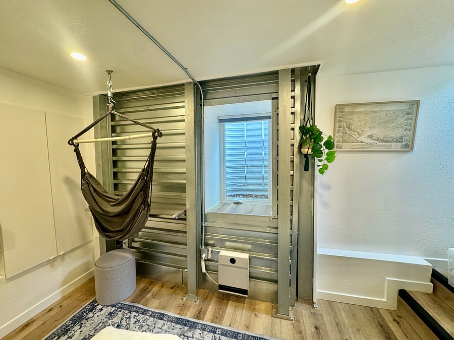 Fold-down stainless steel desk (fixed) for dining/work. Hanging chair and two gray nesting ottomans (one shown) can be repositioned or removed; ottomans function as seating, blanket/storage space, or side tables with a built-in removable wooden tray.
Code-compliant egress window with ladder and blackout blind; tenant-controlled heater below.
Artwork and plants can be stored upon request. Twin XL bed and desk cannot be moved.