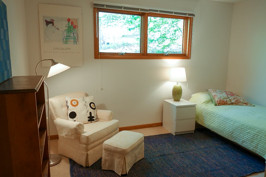 The bedroom is neatly furnished with a comfy chair and footstool for reading, 3 lamps, a bookshelf, and a single bed.