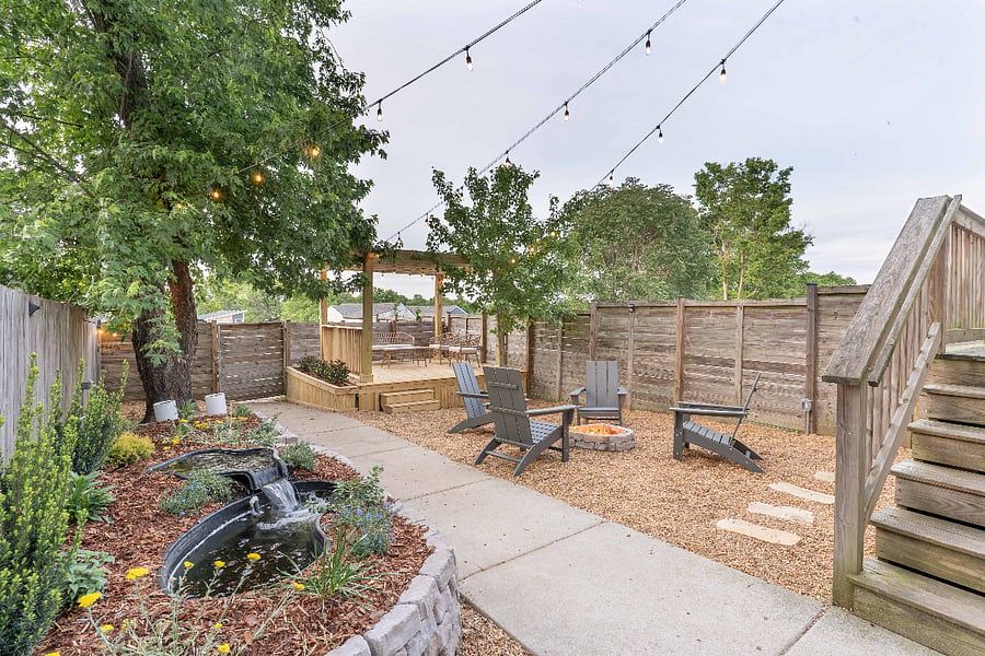 Fenced backyard with seating area, pond, and fire pit. Somewhat private from the neighbors thanks to the trees but still visible on either side